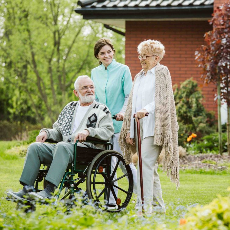 Photo d'un homme âgée dans une chaise roulante dans un jardin. Une femme âgée le suis derrière et une professionnelle pousse la chaise roulante.